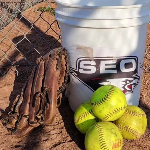 A ball bucket with the SEO logo, a worn glove and a pile of softballs on the infield dirt.